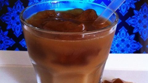 Ein Glas Eiskaffee steht auf einem hellen Untergrund, gefüllt mit gefrorenem Kaffee und einem Strohhalm, vor einem bunten Hintergrund.