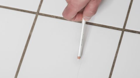 Ein Hand hält einen weißen Kajalstift über einer weißen Wandfliese, um Haarrisse in der Fuge zu kaschieren.