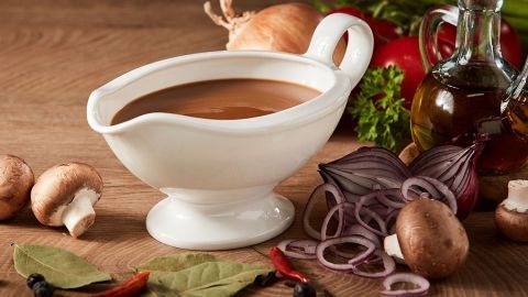 Eine weiße Saucierkanne steht auf einem Holzbrett, gefüllt mit dunkler Sauce, umgeben von Pilzen und frischem Gemüse.