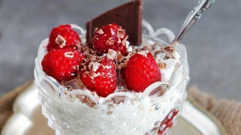 Eine kristallene Schüssel enthält eine cremige Stracciatellacreme, garniert mit frischen Himbeeren und einem Stück Schokolade.