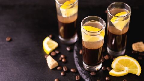 Drei Gläser mit einem dunklen Getränk und Zitronenscheiben stehen auf einem schwarzen Hintergrund, umgeben von Kaffeebohnen.