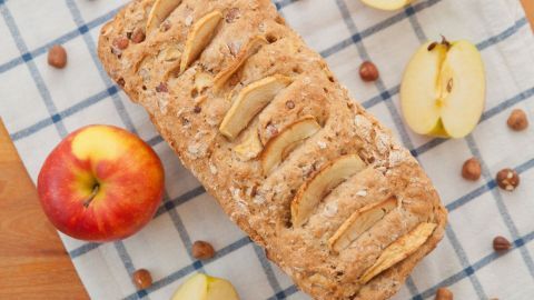 Ein saftiges Apfelbrot liegt auf einem karierten Servietten, umgeben von frischen Äpfeln und Haselnüssen.