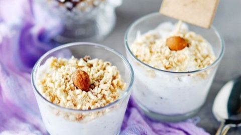 Zwei Dessertgläser mit crunchy Toppings stehen auf einem lila Tuch, umgeben von Nüssen und einem dekorativen Schälchen.