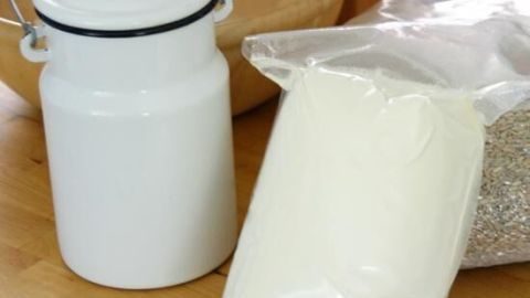 Ein weißes Glasgefäß steht neben einer Tüte mit milchähnlichem Inhalt und einem Sack mit Getreide auf einem Holztisch.