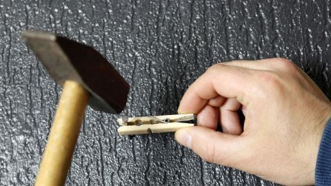 Ein Hammer schlägt auf einen Nägel, der in eine Wand gesteckt ist, während eine Hand eine Klammer hält.