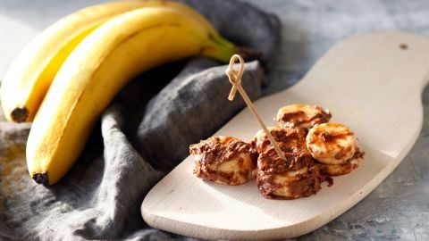 Auf einem Holzbrett liegen mit Schokolade überzogene Bananenscheiben neben frischen Bananen, umgeben von einem grauen Tuch.