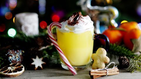 Ein gelbes Getränk mit Sahnehaube und Zuckerstange steht dekorativ zwischen Weihnachtsbaumlichtern und winterlichen Leckereien.