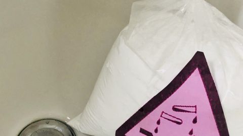 Eine weiße Tüte mit einem Warnschild liegt neben einem verstopften Duschabfluss. Die Tüte enthält wahrscheinlich ein Reinigungsmittel.