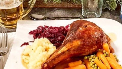 Ein Teller mit einer gekochten Gänsekeule liegt neben Kartoffelbrei, Rotkohl und Karotten, umgeben von dekorativen Kerzen.