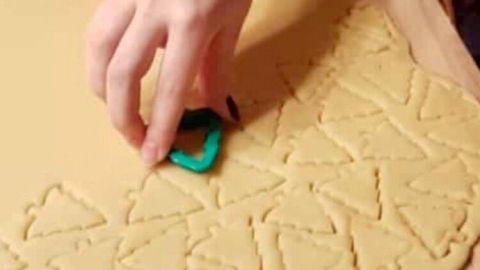 Eine Hand sticht mit einem grünen Ausstecher Weihnachtsplätzchen aus einem Teig auf einer bemehlten Holzplatte aus.