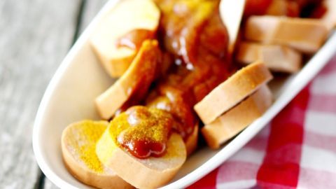 Auf einer Holzoberfläche liegt ein tiefes Geschirr mit Currywurst, umgeben von Scheiben Brötchen und einer roten karierten Serviette.