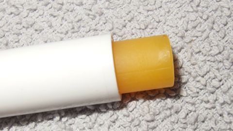 Ein gelber Fettstift liegt auf einem weichen, grauen Stoff. Er ist für die Lippenpflege geeignet und offenbart seine Textur.