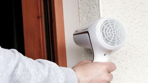 Ein Föhn wird von einer Hand gehalten und bläst warme Luft zu einem Stück Malerkrepp an einer Wand in einem Innenraum.