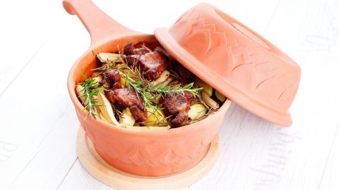 Leckeres Lammfilet mit Kräutern und Gemüse in einem Römertopf auf einem Holzuntersetzer in heller Umgebung.