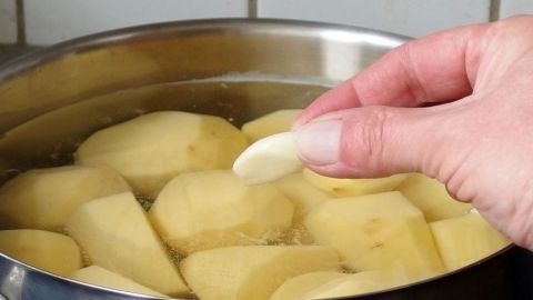 In einem Topf mit kochendem Wasser liegen Salzkartoffeln, während eine Hand eine Knoblauchzehe hinzufügt, um den Geschmack zu verbessern.