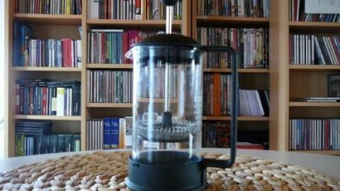 Eine Kaffeepresse steht auf einem runden, geflochtenen Untersetzer vor einem Hintergrund aus Bücher- und CD-Regalen.