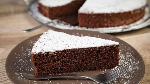 Ein Stück Kuchen liegt auf einem braunen Teller, dekoriert mit Puderzucker, während im Hintergrund weitere Kuchenstücke sichtbar sind.