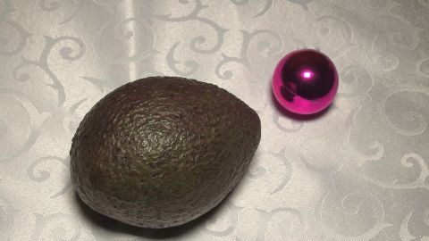 Eine Reife Avocado liegt neben einer pinken Weihnachtskugel auf einem eleganten, glänzenden Tischtuch.