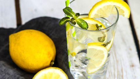 Ein Glas Limonade mit einem grünen Strohhalm steht auf einem Tisch, umgeben von frischen Zitronen und einem grauen Tuch.