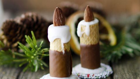 Zwei essbare Kerzen aus Schokolade stehen auf einem Holztisch, umgeben von grünen Tannenzweigen und einer rustikalen Weihnachtsdekoration.
