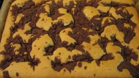 Ein rechteckiger Wolkenkuchen liegt frisch gebacken auf Backpapier, zeigt schokoladige Flecken auf fluffiger, goldener Oberfläche.