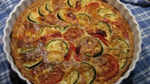 Eine goldbraune Pfannen-Pizza mit Zucchini und Paprika liegt appetitlich in einer weißen Auflaufform auf einem karoblauen Tuch.