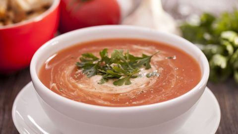 Eine cremige Tomatensuppe steht in einer weißen Schüssel. Frischkäse und Kräuter verleihen ihr ein aromatisches Aussehen.