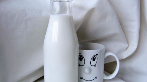 Eine Milchflasche steht neben einer Tasse mit einem lächelnden Gesicht, auf einem hellen Stoffhintergrund.
