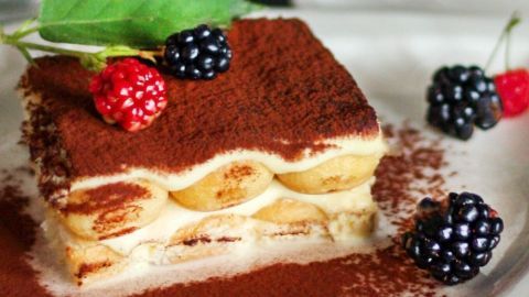 Ein Stück Tiramisu steht auf einem Teller, garniert mit Beeren und einem Blatt, umgeben von Kakaopulver.