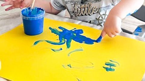 Ein Kind malt mit blauer Farbe auf ein gelbes Blatt, während ein gefüllter Becher Farbe neben ihm steht.