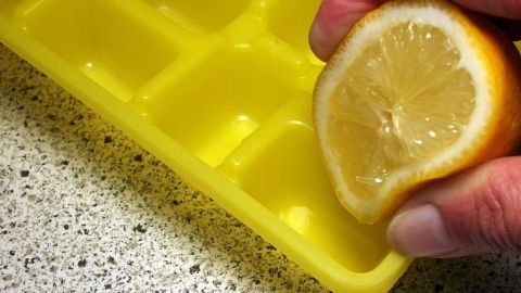 Eine Hand drückt eine halbe Zitrone über einer gelben Eisschale, um frischen Zitronensaft für Eis gegen Halsschmerzen zu gewinnen.