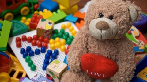 Ein Teddybär mit einem roten Herz sitzt auf einer bunten Spielzeugunterlage, umgeben von vielfältigem, gebrauchtem Spielzeug.