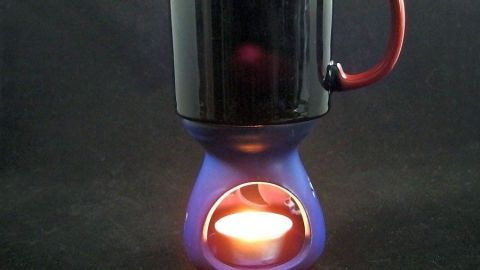 Ein Kaffeebecher steht auf einem Tassenwärmer ohne Strom, der durch eine Kerze beheizt wird, in dunklem Umfeld.