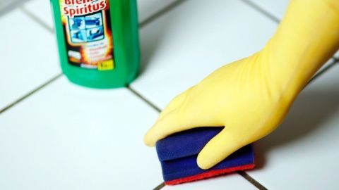 Ein gelber Handschuh hält einen Schwamm, der Zementreste von weißen Fliesen mit Spiritus entfernt, in einer sauberen Umgebung.