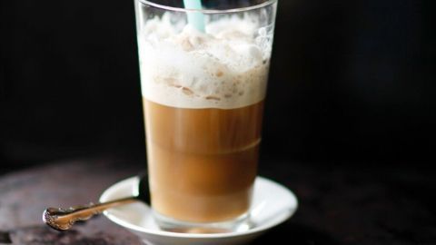 Ein Glas griechischer Frappe steht auf einem Unterteller, gefüllt mit schaumiger Kaffeecreme und einem blauen Strohhalm.