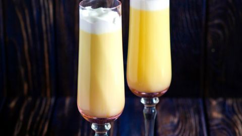 Zwei elegante Gläser mit einem gelben Cocktail, geschichtet, auf einer dunklen Holzoberfläche, mit einer Sahnehaube dekoriert.