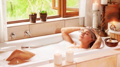 Bei Husten ist es wichtig, die Atemwege feucht zu halten – dies ist besonders gut mit einem heißen Bad zu erreichen Eine Frau entspannt in einer Badewanne mit Wasser, umgeben von Kerzen und Pflanzen, während sie Husten lindert.