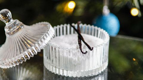 Ein elegantes Glasgefäß steht offen auf einem Tisch, gefüllt mit Zucker und dekoriert mit Vanilleschoten, im festlichen Umfeld.