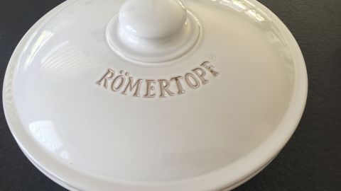 Ein Römertopf eignet sich hervorragend als Brottopf und hält dein Brot länger frisch. Die unglasierte Innenseite reguliert Feuchtigkeit, verhindert Schimmel und sorgt für ein perfektes Aroma. Ideal für deine Backwaren. Ein weißer Römertopf mit Deckel liegt auf einer dunklen Oberfläche. Der Schriftzug "RÖMERTOPF" ist auf dem Deckel eingraviert.