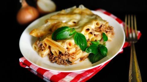 Ein Teller Rigatoni al Forno liegt auf einer rot-weiß karierten Serviette. Saftige Pasta mit Fleischsoße und frischem Basilikum wird serviert.
