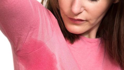 Frau betrachtet besorgt Deoflecken auf farbigem T-Shirt, hebt Arm, zeigt Verfärbung unter der Achsel in heller Umgebung.