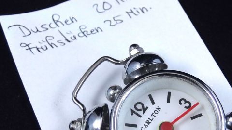 Eine Uhr liegt neben einem Zettel, auf dem steht: "Duschen 20 Min., Frühstücken 25 Min." Zeit messen für den Morgen.