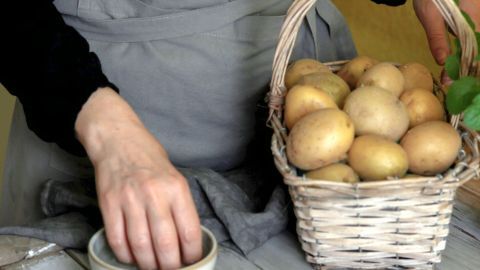 Eine Hand greift nach einer Kartoffel aus einem Korb, während die andere Hand eine Schale mit etwas Inhalt hält.
