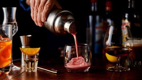 Ein Barkeeper gießt einen rosafarbenen Cocktail in ein Glas, umgeben von verschiedenen Spirituosen und Zutaten.