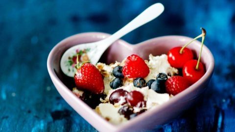 Eine herzförmige Schale mit cremigem Joghurt wird mit frischen Beeren und Kirschen dekoriert, auf einem blauen Hintergrund.
