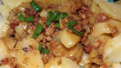 Ein warmer Kartoffelsalat wird appetitlich präsentiert, mit Kartoffeln und Speckstücken, garniert mit frischem Schnittlauch.
