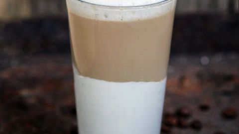 Ein Glas mit schaumiger Milch ist gefüllt, die obere Schicht ist hellbraun und die untere weiß, umgeben von Kaffeebohnen.