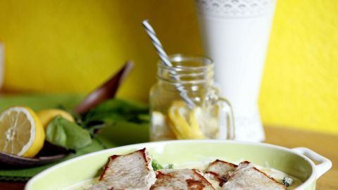 Braten in einer Auflaufform auf Holztisch mit Zitrone und Glas im Hintergrund, neben gelber Wand und weißer Vase.