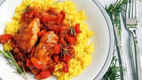 Ein Teller mit Hähnchen in einem bunten Curry, serviert auf gelben Reis, umgeben von frischen Kräutern.