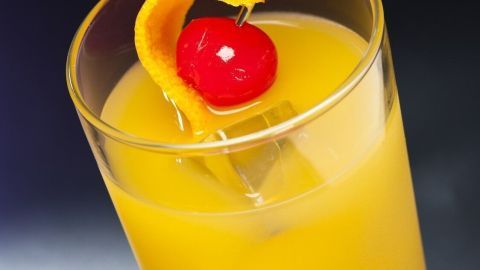 Ein Glas mit Schlammbowle steht auf einem dunklen Untergrund, gefüllt mit einer gelben, fruchtigen Flüssigkeit und dekoriert mit einer Kirsche und einer Orangenscheibe.